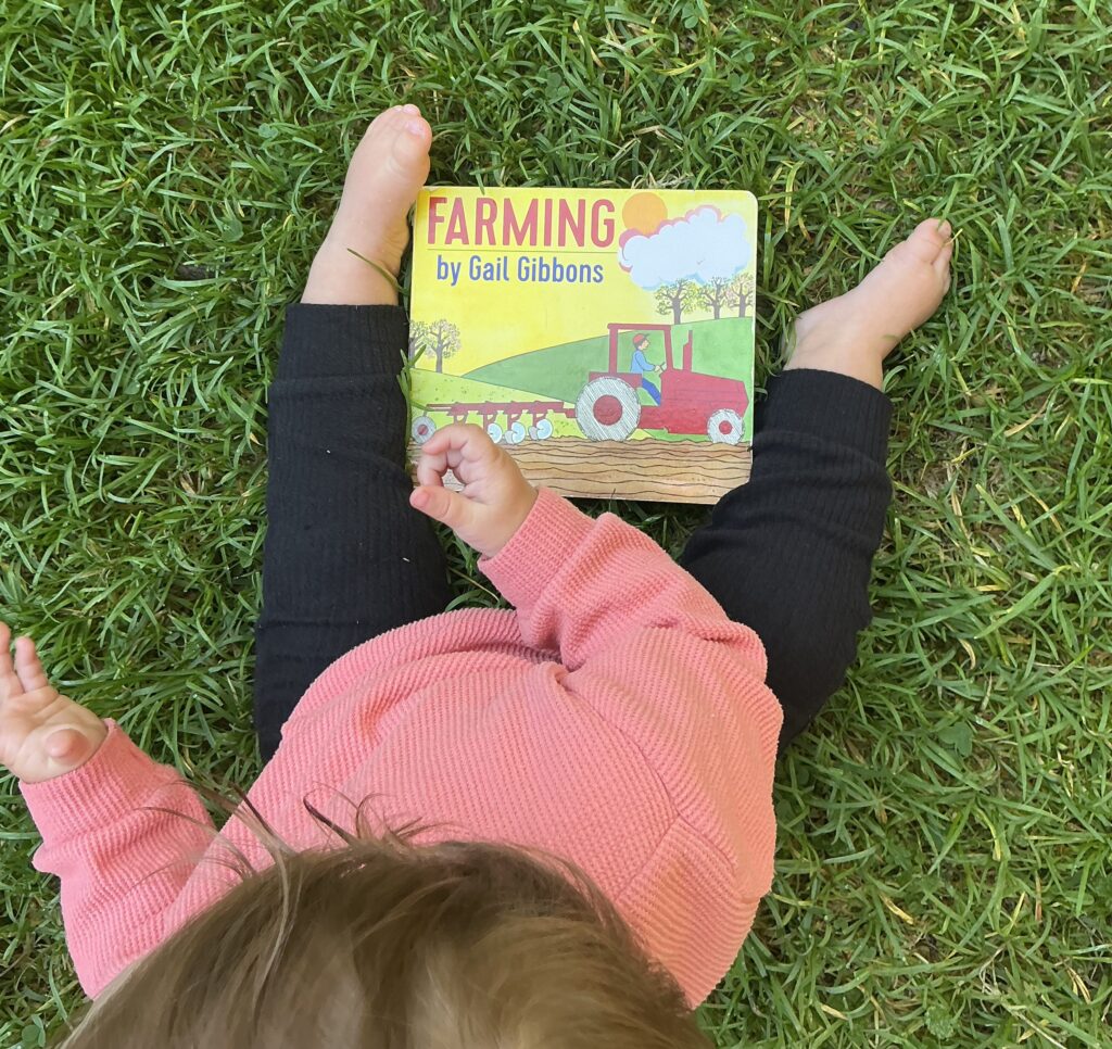 Baby playing in the grass with a board book about farming